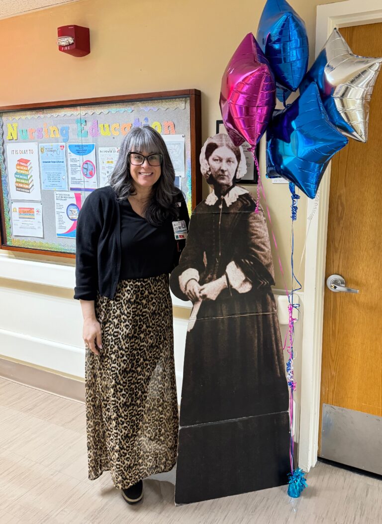 Kristina standing next to a Florence Nightingale cutout in a hospital hallway next to the Nursing Education bulletin board