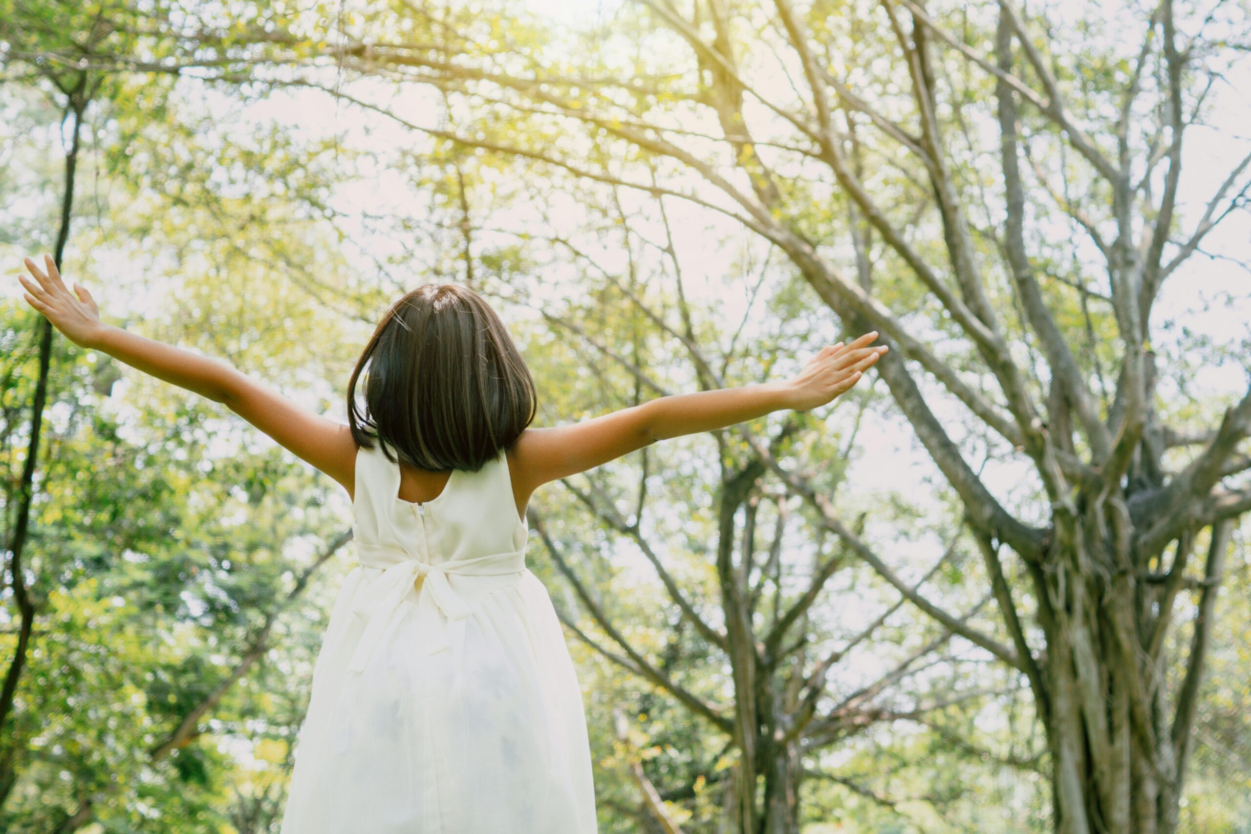 A girl in a white dress with her arms spread wide under sunlit trees